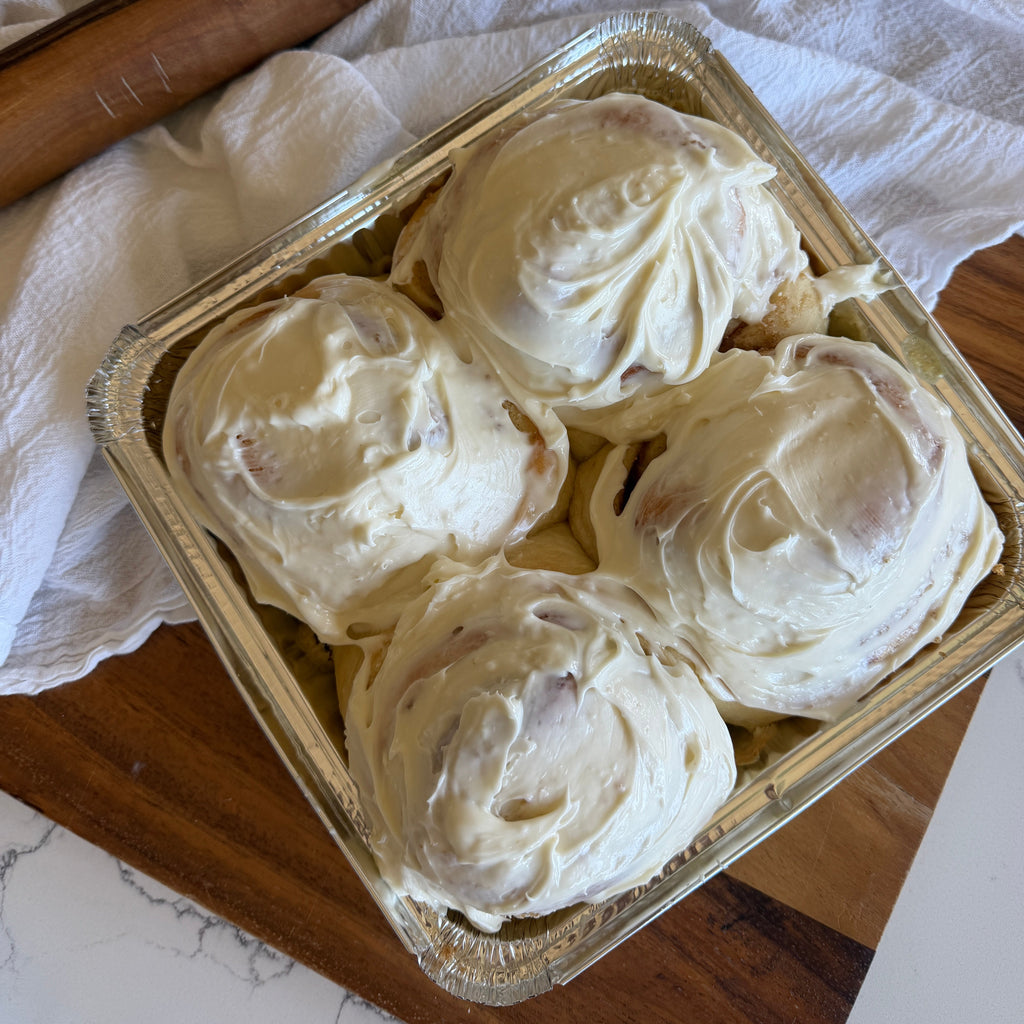 Four frosted cinnamon rolls in a square aluminum pan, styled on a wooden cutting board with a white cloth and a wooden rolling pin.